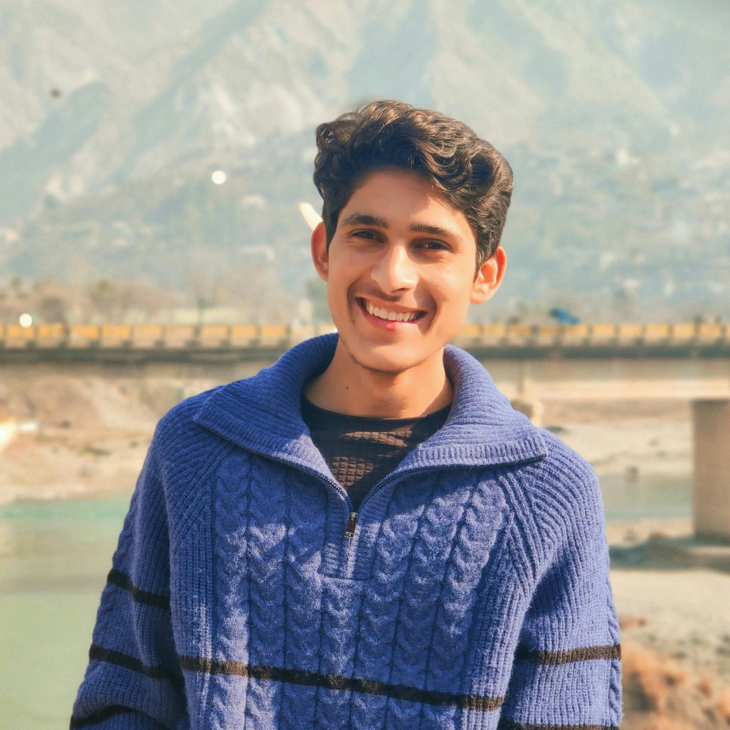A cheerful young man in a blue sweater stands outdoors with mountains in the background.